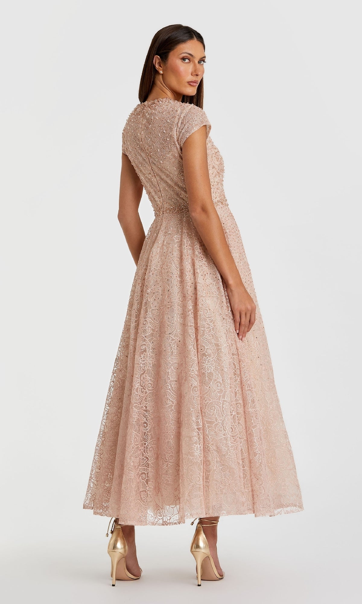 Woman in a pale pink Mac Duggal 20371 Cap Sleeve A-Line Formal Dress, featuring lace details and short sleeves, stands in profile looking over her shoulder, paired with gold high heels against a plain background.